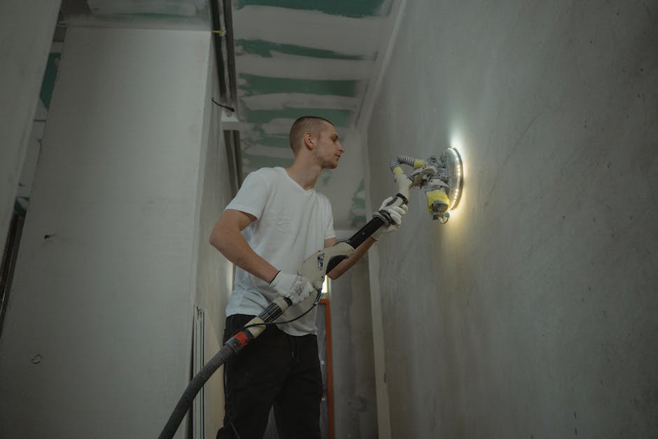 A man sanding an interior wall with an automatic sanding tool during renovation work indoors.